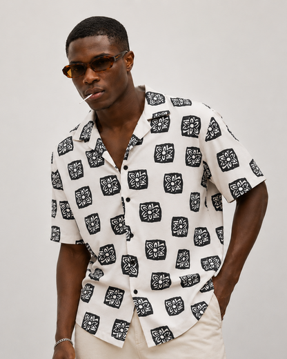 Man wearing a white shirt with black pattern and sunglasses on a plain background